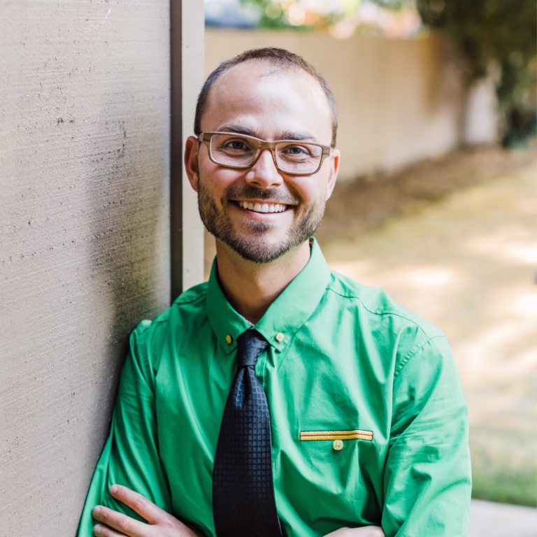 Smiling man with glasses in a bright green shirt and dark tie, leaning against a beige wall outdoors.