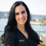 Smiling woman with long black hair in a black top with embroidered text, outdoors by water.