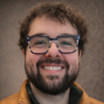 Smiling man with dark hair and a beard wearing striped blue glasses and a brown jacket (close-up). Dim background.