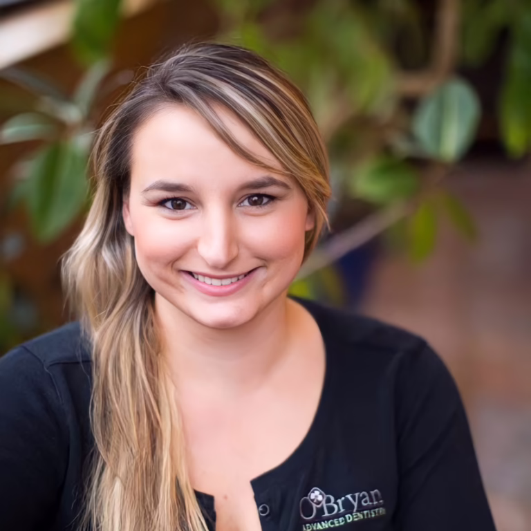 Smiling woman with long blonde hair wearing a black polo with a dental logo, indoors with greenery in the background.