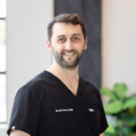 Smiling male healthcare professional in black scrubs standing in a bright indoor space, possibly clinic or hospital.