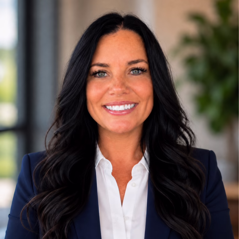Professional headshot: smiling woman with long dark hair, wearing a navy blazer and white shirt in an office setting.