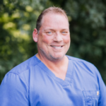 Smiling man in blue scrubs outdoors with a blurred green leafy background, facing camera.