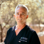 Smiling dentist in black scrubs outdoors with a blurred natural background and Elevation Dental Group logo on the chest pocket.