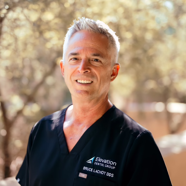 Smiling dentist in black scrubs outdoors with a blurred natural background and Elevation Dental Group logo on the chest pocket.