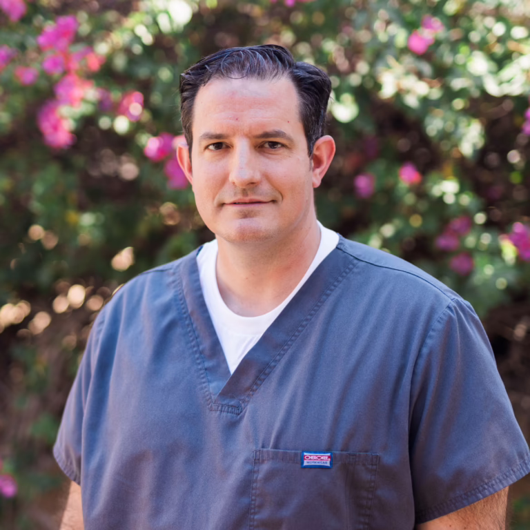 Portrait of a male healthcare worker in gray scrubs outdoors with pink flowers in the background.