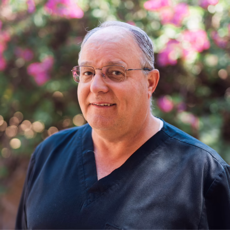 Portrait of a smiling man wearing dark scrubs and glasses, outdoors with pink flowers in the background.