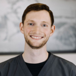 Portrait of a smiling man in gray scrubs with a neutral blurred background.