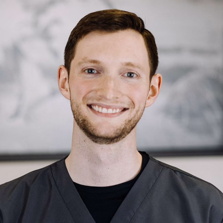 Portrait of a smiling man in gray scrubs with a neutral blurred background.