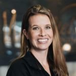 Professional headshot of a smiling woman with long light brown hair, wearing a black top, in a blurred office setting.