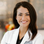 Smiling female doctor in a white coat with name badge, facing the camera in a clinic setting.