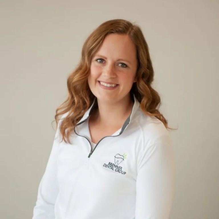Portrait of a smiling woman with wavy red hair wearing a white polo with Brenner Dental Group logo.