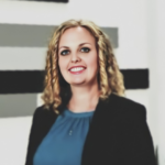 Professional woman with blonde wavy hair, wearing a teal blouse and black blazer, smiling at the camera with abstract wall art in the background.