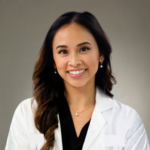 Smiling woman in a white lab coat with black top, wearing pearl earrings and a small pendant necklace, portrait-style.