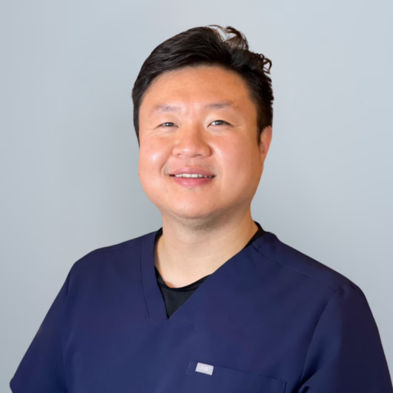 Smiling healthcare professional in navy scrubs against a light gray background, looking at the camera.