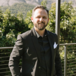 Smiling man in a black suit with a light blue pocket square, standing outdoors near a railing against green foliage.
