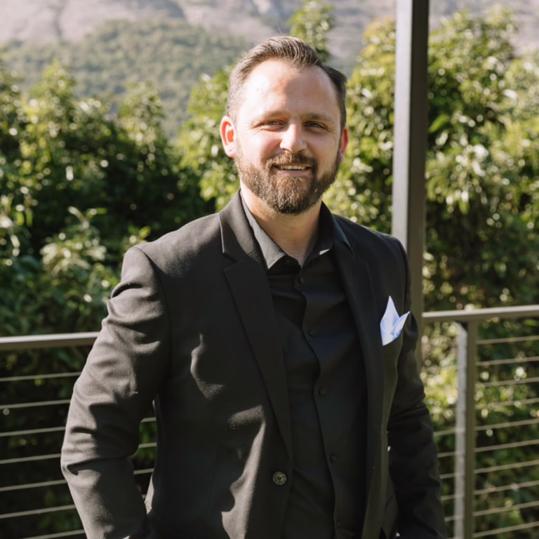 Smiling man in a black suit with a light blue pocket square, standing outdoors near a railing against green foliage.