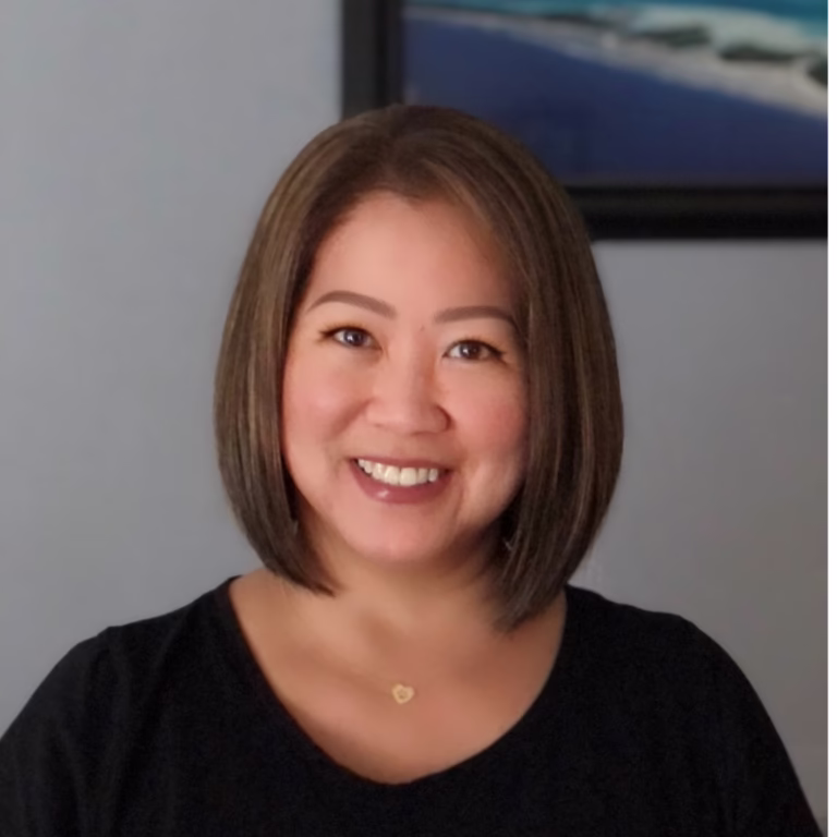 Smiling woman with a short brown bob, wearing a black top and a small heart necklace indoors against a gray wall.