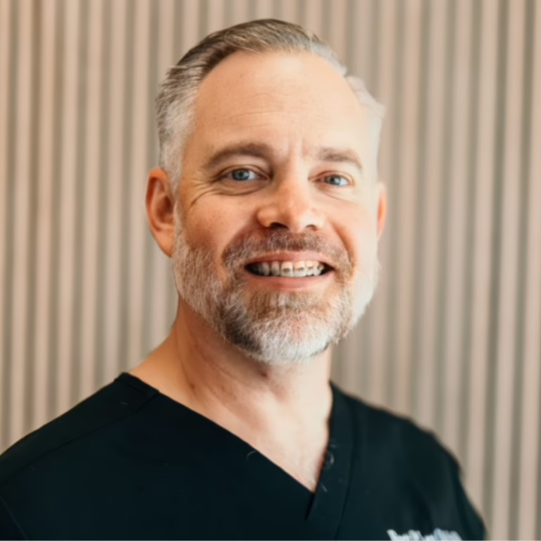 Smiling middle-aged man with gray beard wearing black scrubs against a striped wall.
