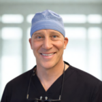 Smiling male surgeon in a blue surgical cap and black scrubs, with glasses hanging from his neck, in a bright clinic setting