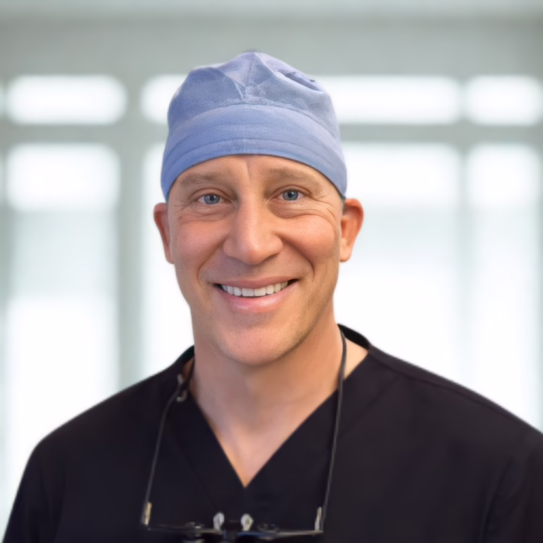 Smiling male surgeon in a blue surgical cap and black scrubs, with glasses hanging from his neck, in a bright clinic setting