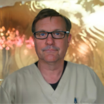 Man in light medical scrubs with black glasses looking at the camera, a soft gold blurred background behind him.