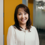 Smiling woman in a white blouse standing beside a yellow wall in an office corridor.