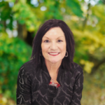 Head-and-shoulders portrait of a smiling woman with dark hair, wearing a black patterned jacket, outdoors among green foliage, with hoop earrings and a pendant.