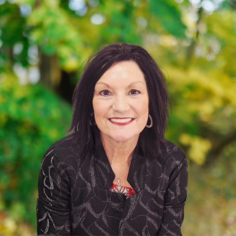 Head-and-shoulders portrait of a smiling woman with dark hair, wearing a black patterned jacket, outdoors among green foliage, with hoop earrings and a pendant.