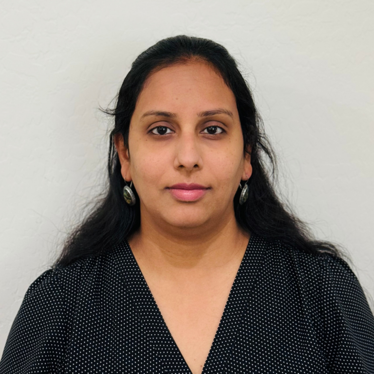 Portrait of a woman with long dark hair, wearing a black polka-dot blouse and silver earrings, facing the camera against a light wall.