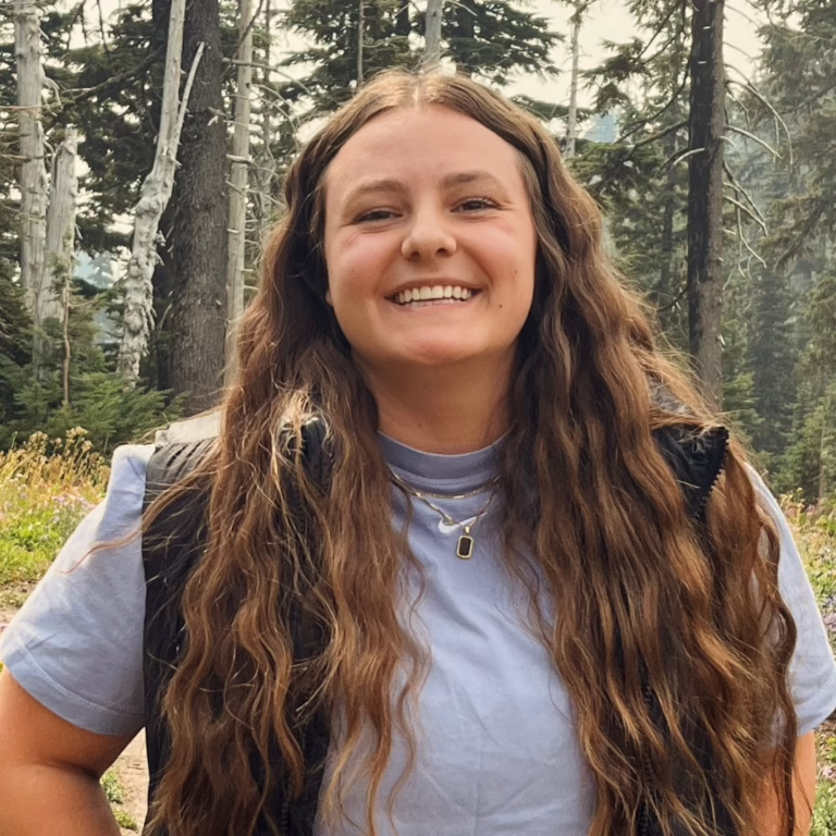 Smiling young woman with long wavy hair in a forest, wearing a light blue shirt and black vest.