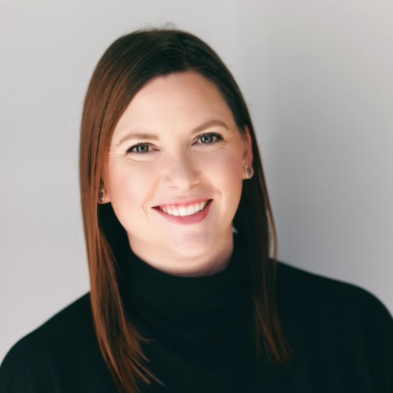 Portrait of a smiling woman with brown hair wearing a black turtleneck against a light gray background, looking at the camera.