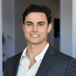 Professional headshot of a smiling man in a dark suit and white shirt, neutral office background.