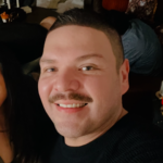 Close-up portrait of a man with short dark hair and a mustache smiling at the camera, wearing a black sweater/tee in an indoor setting.