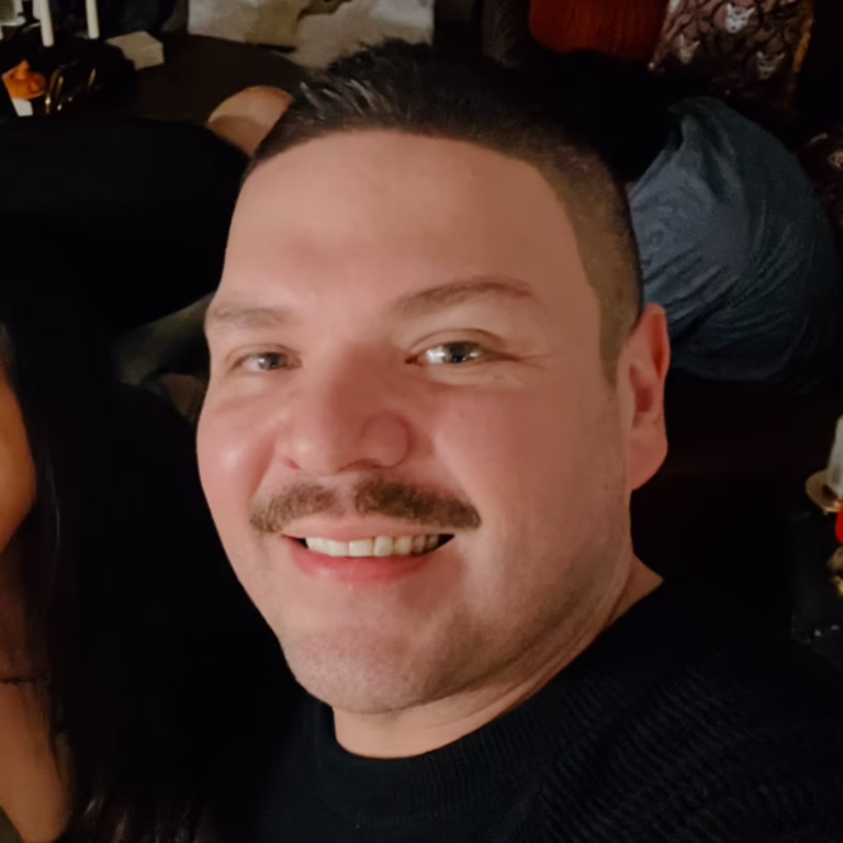 Close-up portrait of a man with short dark hair and a mustache smiling at the camera, wearing a black sweater/tee in an indoor setting.
