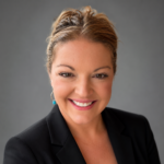 Portrait of a smiling woman with blond hair tied back, wearing a black blazer, against a gray studio backdrop.