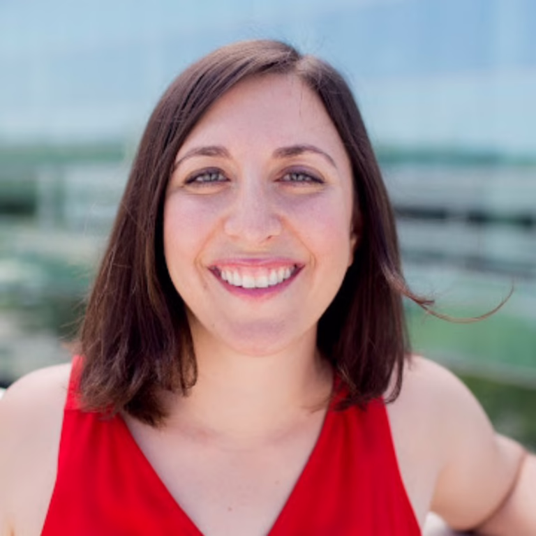 Portrait of a smiling woman with shoulder-length brown hair wearing a red sleeveless top outdoors. (Informative)