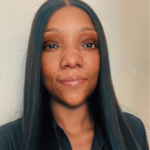Portrait of a smiling woman with long straight black hair wearing a dark shirt, against a beige wall background.