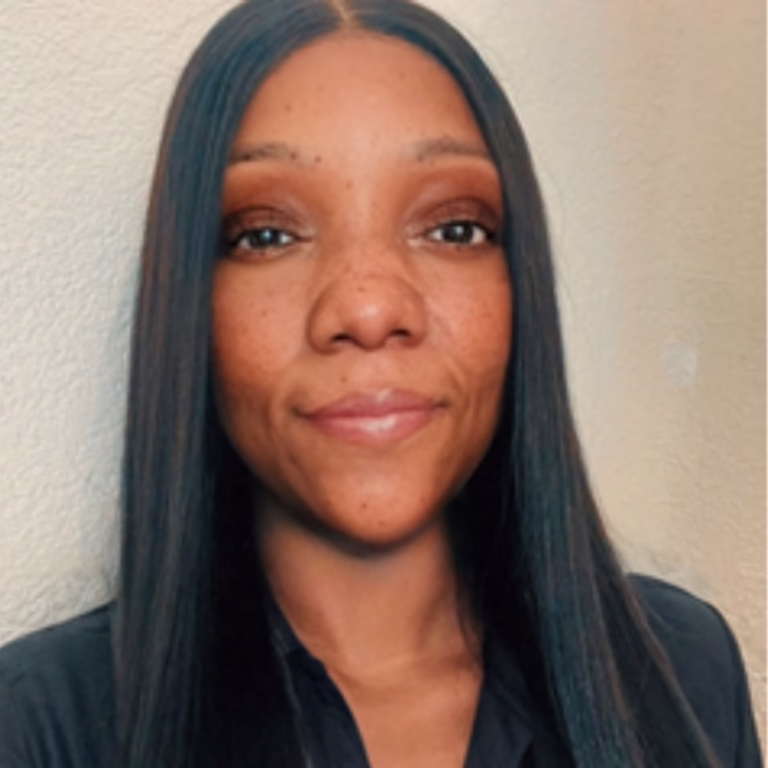 Portrait of a smiling woman with long straight black hair wearing a dark shirt, against a beige wall background.