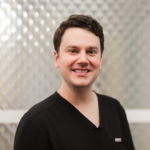 Smiling man with short dark hair in black scrubs, standing in front of a textured metallic background.