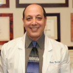 Smiling male doctor in a white coat with a gray shirt and blue tie, standing in a certificate-lined office.