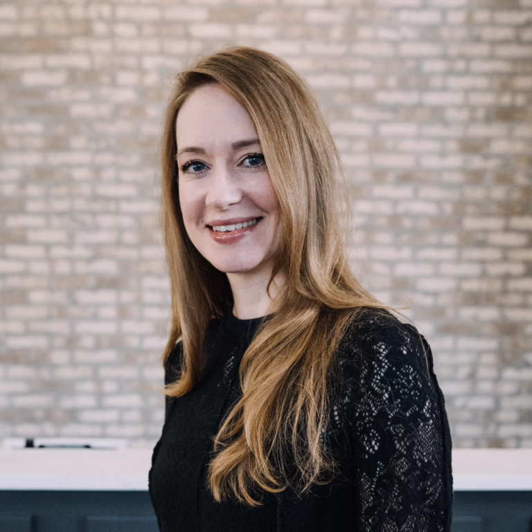 Head-and-shoulders portrait of a smiling woman with long blonde hair in a black lace top, posed in front of a light brick wall background.