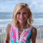 Smiling blonde woman in a colorful floral sleeveless dress with white circular trim, standing on a balcony overlooking the ocean.