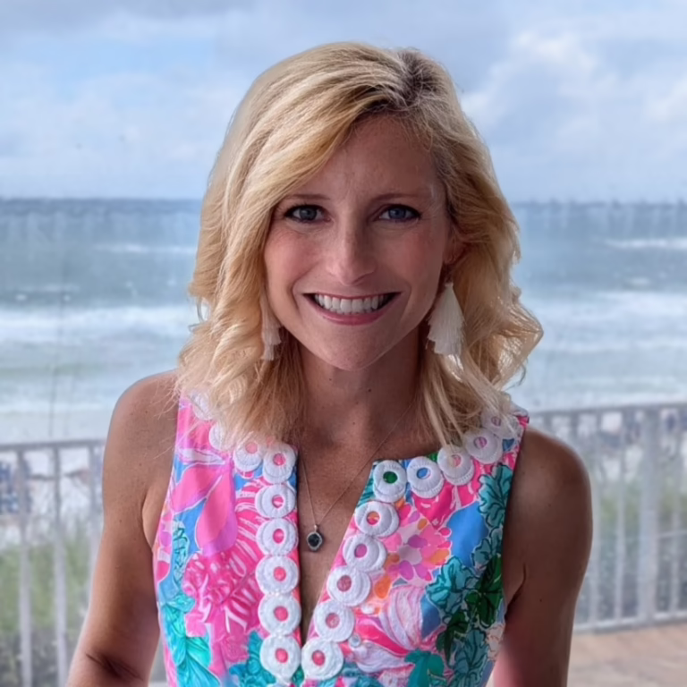 Smiling blonde woman in a colorful floral sleeveless dress with white circular trim, standing on a balcony overlooking the ocean.