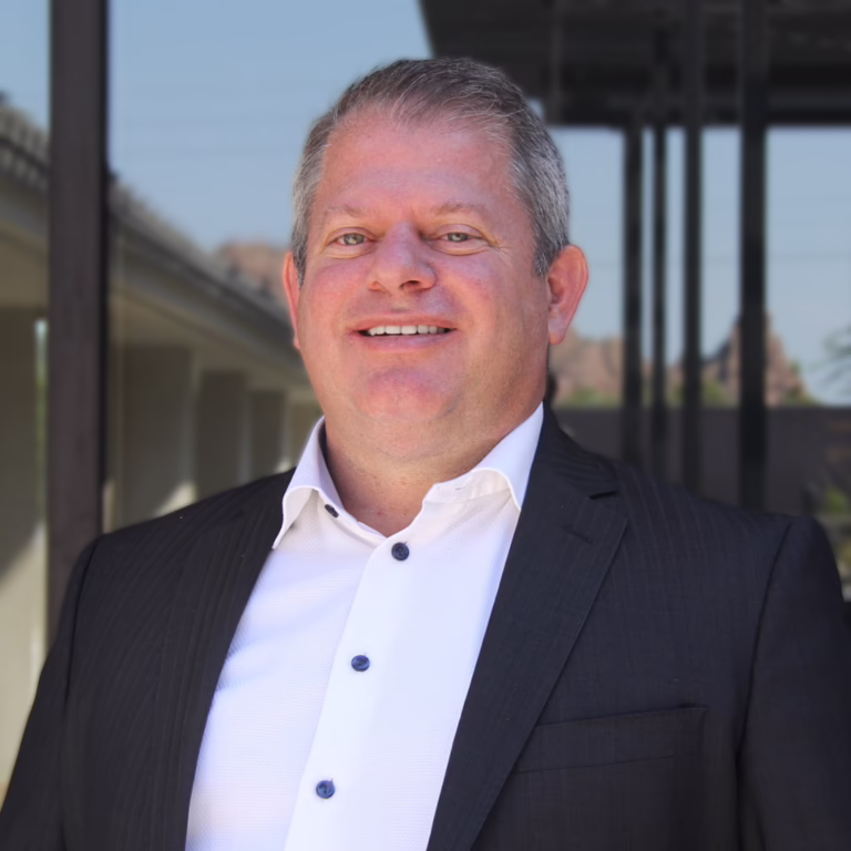 Head-and-shoulders portrait of a smiling man in a dark suit and white shirt outdoors beside a building.