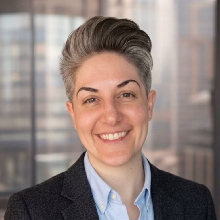 Smiling professional portrait of a person with short gray hair in a blazer and light blue shirt, office background.