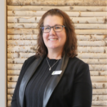 Smiling woman with shoulder-length brown hair and glasses, wearing a black blazer and name badge, in front of a light wooden paneled wall.
