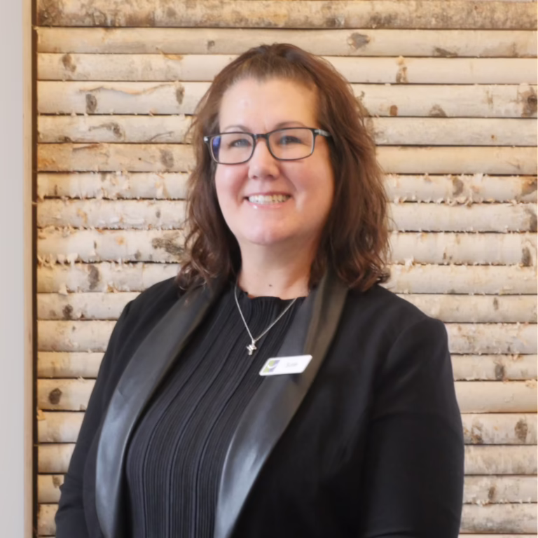 Smiling woman with shoulder-length brown hair and glasses, wearing a black blazer and name badge, in front of a light wooden paneled wall.