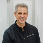 Smiling man in a dark zip-up jacket with 'Smile Moore Dentistry' embroidery, portrait in a clinical setting.