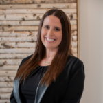 Smiling woman in a black blouse with leather trim, posing for a professional headshot in front of a wooden paneled wall.